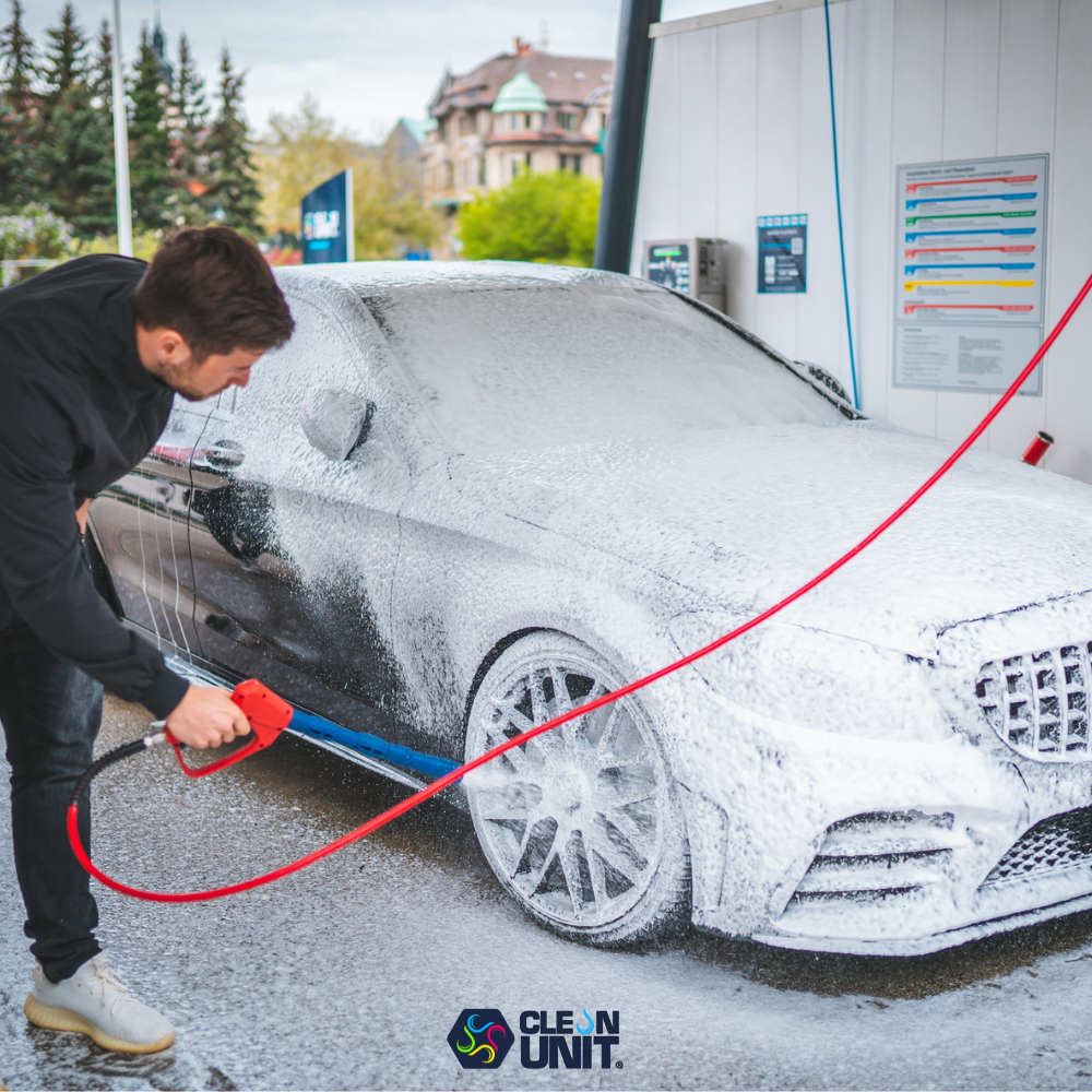 Mann reinigt mit Hochdrucklanze ein Auto in einer SB-Waschanlage von Clean Unit. Das Fahrzeug ist vollständig mit Powerschaum bedeckt. Moderne Waschbox mit klarer Beschilderung im Hintergrund.