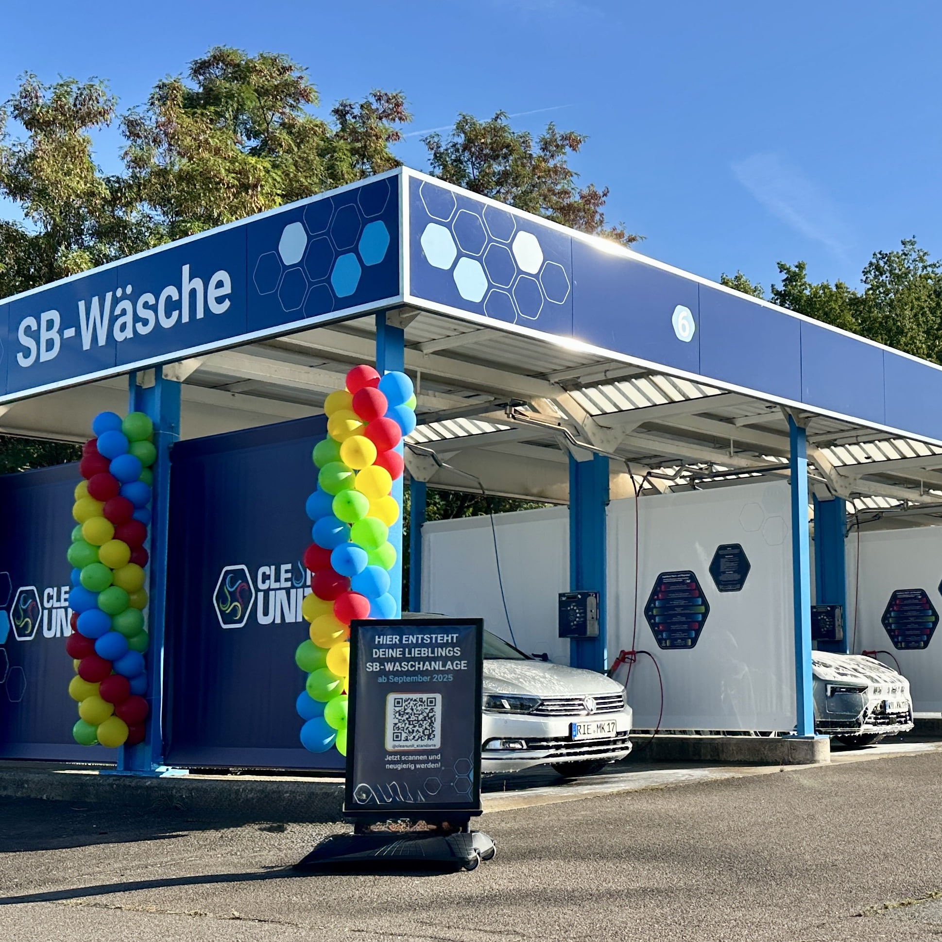 Moderne SB-Waschanlage von Clean Unit mit blauen Wänden und bunten Luftballons zur Eröffnung. Mehrere Waschboxen, Autos in der Anlage und ein Aufsteller mit QR-Code im Vordergrund.